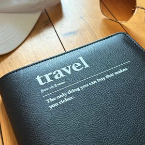 Pode incluir: Um diário de viagem de couro preto com a palavra "travel" em texto branco e a definição "[trav-uh-l] substantivo A única coisa que você pode comprar que o torna mais rico."