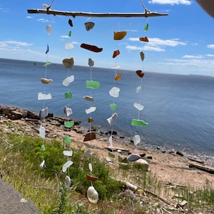 May include: A sea glass wind chime with a wooden dowel and white string hanging from it. The wind chime is made of various colors of sea glass, including green, blue, white, and brown. The wind chime is hanging in front of a body of water.