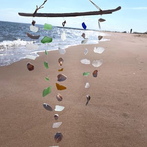 May include: A handmade wind chime crafted from driftwood and sea glass. The chime features various shapes and colors of sea glass, including green, white, and amber, strung together with twine. The background shows a beach and ocean.