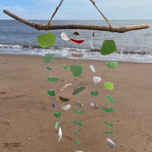 May include: A sea glass and shell wind chime hanging from a driftwood branch. The chime features various shades of green sea glass, white shells, and a few red and brown pieces. The chime is hanging against a sandy beach background with blue sky and ocean in the distance.