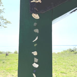 May include: A vertical hanging mobile composed of a series of small, smooth sea glass pieces and white seashells strung together on a thin cord. The collection includes translucent white, green, and blue glass fragments alongside various shell shapes.