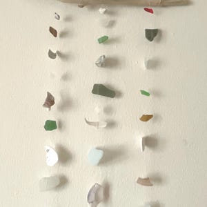May include: A wall hanging featuring a horizontal piece of driftwood suspended by a twine cord with a small blue bead. Three vertical strands hang from the wood, each decorated with various pieces of multi-colored sea glass and small white shells.