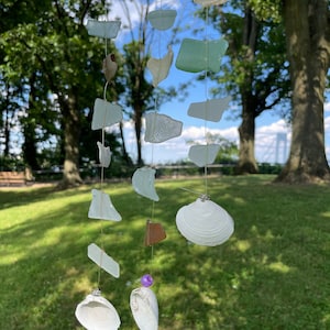 May include: A sea glass and seashell wind chime hanging from a tree branch. The chime features various shapes and sizes of sea glass in shades of green, blue, and white, along with white seashells.