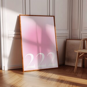 以下が含まれることがあります： ピンクのグラデーションと白い数字「222」が入った額入りのアートプリント。木製の床と白い壁のある部屋に飾られており、壁に立てかけられています。