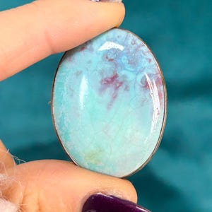 May include: Oval-shaped brooch with a silver-colored metal frame. The brooch features a marbled design in shades of turquoise and maroon. The surface has a cracked, aged appearance.