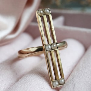 May include: A gold ring with a rectangular design featuring three horizontal bars and five small white pearls. The ring is set on a pink fabric background.