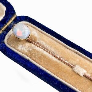 Op de afbeelding: Een antieke opaalspeld in een blauwe fluwelen doos. De speld heeft een gouden zetting rond de ovale opaal edelsteen. De steel van de speld heeft een gedraaid ontwerp. De doos heeft een gouden scharnier en een beige fluwelen interieur.