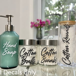 Peut inclure: Quatre contenants de comptoir de salle de bain avec des étiquettes en vinyle noir. Une bouteille à pompe verte étiquetée "Savon pour les mains", un pot en verre transparent étiqueté "Boules de coton", un pot en verre transparent étiqueté "Cotons-tiges", et un pot en verre transparent avec un couvercle en bambou étiqueté "Ronds de coton".