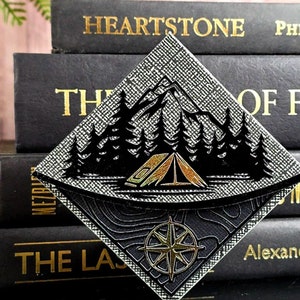 May include: A black and gold diamond-shaped bookmark with a design of a tent in a forest with mountains in the background. The bookmark also features a compass design.