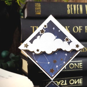 May include: A blue and white paper bookmark featuring a white cloud and gold glitter stars. The bookmark is diamond-shaped and is positioned on top of a stack of books.