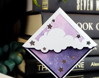May include: A purple and white paper bookmark with a cloud and stars design. The bookmark is shaped like a diamond and has a glittery finish.