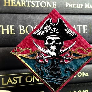May include: A diamond-shaped patch featuring a black pirate skull wearing a tricorn hat with a skull and crossbones emblem. Below the skull are two crossed gold swords over a dark teal scene of a sailing ship on wavy water with a sea creature.