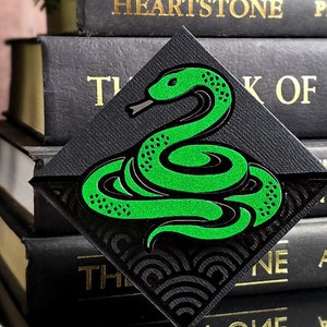 May include: A black and green bookmark with a snake design. The snake is curled up and has a green body with black spots. The bookmark is shaped like a triangle and has a black background.