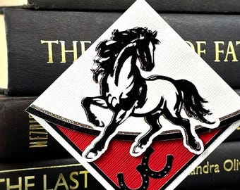 Może przedstawiać: Naszywka w kształcie rombu z czarno-białą ilustracją stającego dęba konia. Pod koniem znajduje się czarna ikona podkowy na czerwonym, teksturowanym tle. Naszywka ma białą obwódkę i jest umieszczona na czarnych książkach.