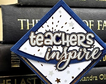 Może przedstawiać: Tabliczka w kształcie rombu z białym tłem i napisem "teachers inspire" wykonanym srebrnym brokatem. Tabliczka ma ciemnoniebieską obwódkę i jest ozdobiona małymi srebrnymi i złotymi gwiazdkami oraz złotymi akcentami przypominającymi konfetti.