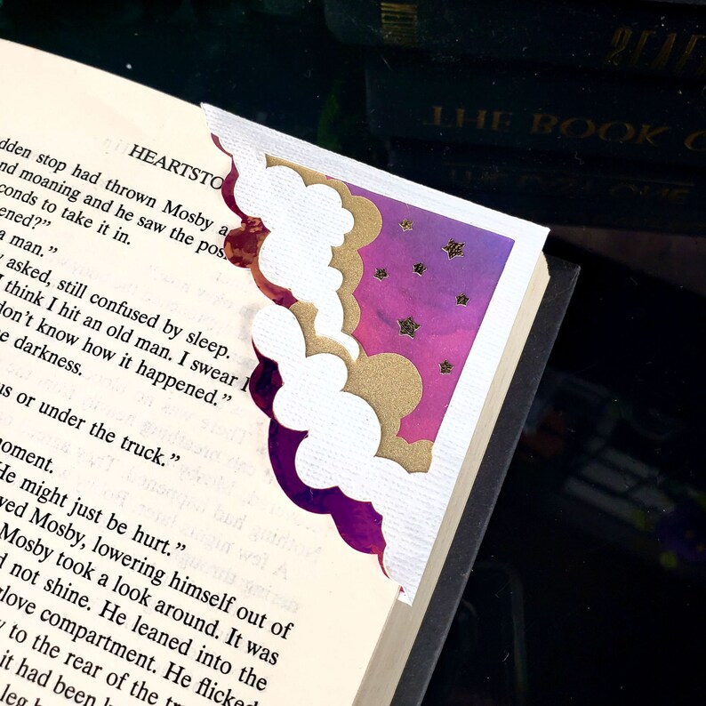 May include: A corner bookmark featuring layered cloud shapes in white and gold, set against a purple and pink watercolor background with small gold stars. The bookmark is attached to the corner of an open book page with printed black text.