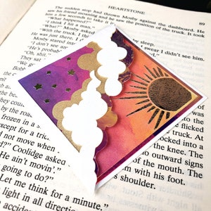 May include: A triangular corner bookmark featuring a split design. One side displays a purple night sky with gold stars, while the other side shows an orange and red sunset with a large gold sun. White die-cut clouds overlap the center seam.