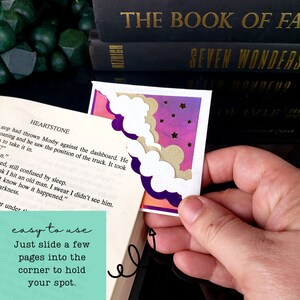 May include: A corner bookmark featuring a layered design with white clouds, a gold glittery cloud, and a purple and pink gradient background with small gold stars. The bookmark is shown attached to the corner of an open book page.