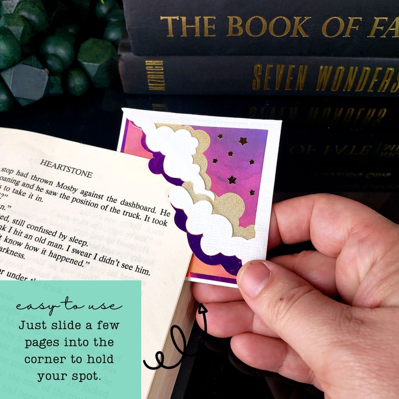 May include: A corner bookmark featuring a layered design with white clouds, a gold glittery cloud, and a purple and pink gradient background with small gold stars. The bookmark is shown attached to the corner of an open book page.