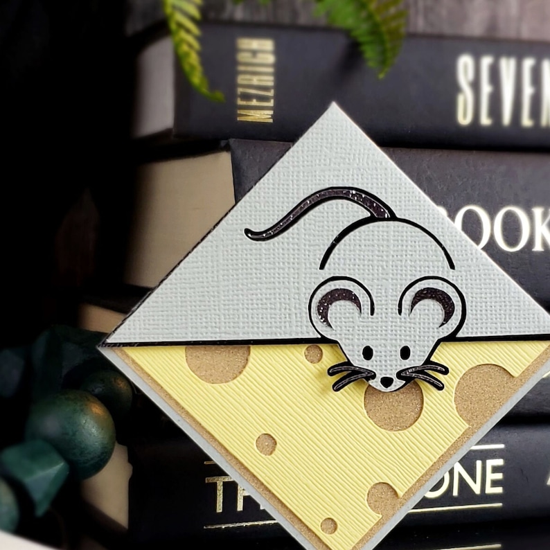 May include: A gray and white mouse bookmark with a cheese design. The mouse is holding a piece of cheese with holes in it.
