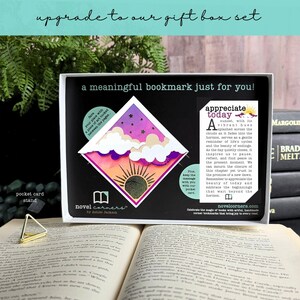 May include: A diamond-shaped bookmark featuring a sunset illustration with purple, orange, and pink gradients, white clouds, and a sun icon. It is presented in a black box with text, including the phrase 'a meaningful bookmark just for you!' and a pocket card stand.