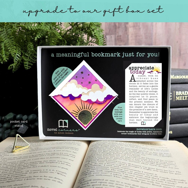 May include: A diamond-shaped bookmark featuring a sunset illustration with purple, orange, and pink gradients, white clouds, and a sun icon. It is presented in a black box with text, including the phrase 'a meaningful bookmark just for you!' and a pocket card stand.