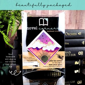 May include: A diamond-shaped bookmark inside a clear plastic pillow box. The bookmark features a purple and orange sunset design with white clouds and a sunburst graphic. Text on the packaging includes 'novel corners' and instructions.