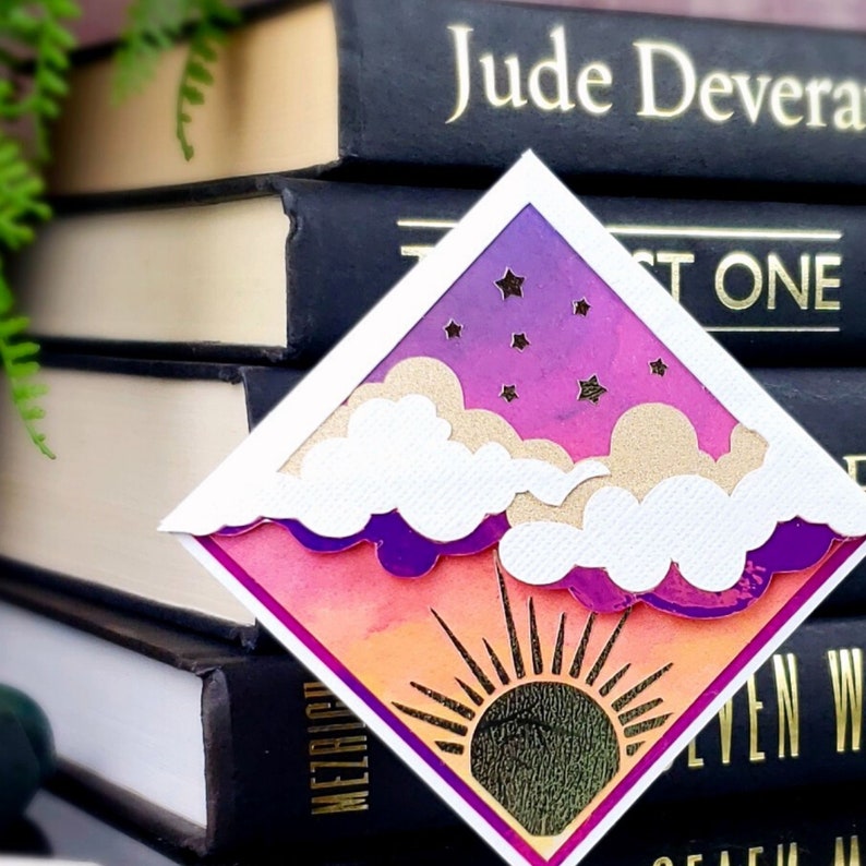 May include: A diamond-shaped paper cutout featuring a sunset scene with an orange and pink gradient sky, a dark sun with radiating black lines, and layered white and gold clouds against a purple starry night sky with small cut-out stars.