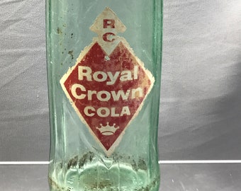 Pode incluir: Garrafa vintage de vidro verde Royal Crown Cola. O rótulo apresenta "Royal Crown COLA" em branco sobre um losango vermelho. A garrafa tem uma forma clássica, ligeiramente curvada.
