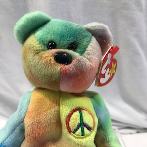 May include: A colorful plush bear toy with a peace sign on its chest. The bear features a rainbow of colors, including green, blue, yellow, and orange. It has black eyes and a red tag attached to its ear.