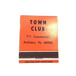 May include: A vintage matchbook with an orange cover. The text "TOWN CLUB" is printed in black, along with the address "711 Commercial, Atchison, Ks. 66002." The bottom reads "CLOSE COVER BEFORE STRIKING MATCH."