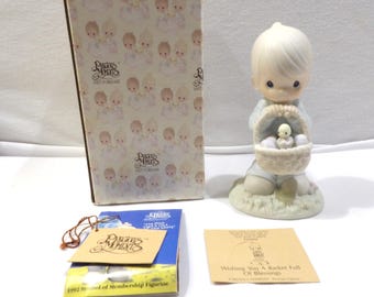 May include: A Precious Moments figurine of a young child holding a basket with a chick and eggs. The figurine is on a grassy base. Includes the original box and tags. The box has a repeating pattern of Precious Moments figures.
