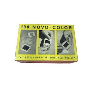 May include: A yellow box with the text "100 NOVO-COLOR" and "2"x"2" METAL COLOR SLIDES SWISS MADE MOD. DÉP.". The box shows three images of hands inserting a metal slide into a projector.