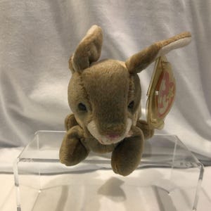 May include: A brown plush rabbit toy with soft fur, long ears, and a small pink nose. The stuffed animal features dark eyes and a light-coloured muzzle area. A clear plastic heart-shaped tag with the letters TY is attached to the rabbit's ear.