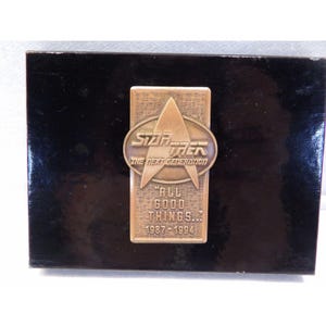 Könnte beinhalten: Rechteckige, bronzefarbene Star Trek: The Next Generation-Plakette mit dem Starfleet-Abzeichen und dem Text "ALL GOOD THINGS... 1987-1994" auf einem schwarzen Holzsockel.