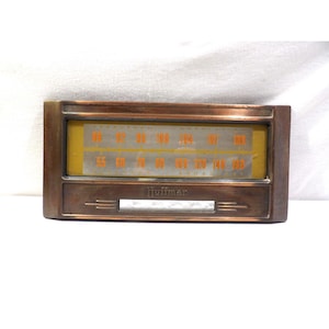 Op de afbeelding: Een rechthoekige Hoffman radio-wijzerplaat met een metalen, bronskleurig frame. Het display heeft een gele achtergrond met oranje frequentiecijfers voor FM- en AM-banden, met de merknaam Hoffman in reliëf onder het glazen paneel.