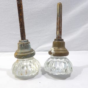 May include: Two antique glass door knobs with brass bases and rusty metal stems. The clear glass knobs have a ribbed design. One stem is heavily rusted, while the other shows less rust. The knobs are set against a white background.