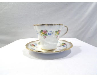 May include: White teacup and saucer set with a floral pattern in pink, yellow, and blue. The edges of the cup and saucer are scalloped and trimmed with gold. The teacup has a delicate handle.