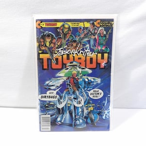 Op de afbeelding: Een stripboek getiteld Toyboy nr. 2 van Continuity Comics. De cover toont een centraal personage in een blauw mechanisch pak, omringd door verschillende figuren. De tekst bevat Jason Kriter, Toyboy en tekstballonnen met de woorden Hey, Dirtbags en Your Victim's Here. De achtergrond is een donkerblauw verloop.