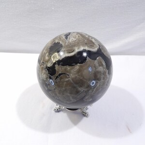 May include: A polished sphere with a marbled appearance, featuring shades of gray, black, and tan. The sphere is displayed on a silver-colored, ornate stand with three feet. The surface of the sphere has a glossy finish.