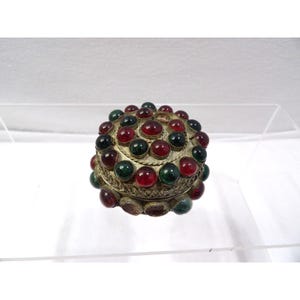 May include: A small, round metal trinket box featuring an intricate, textured surface adorned with numerous cabochon-style stones in alternating deep red and dark green colors, arranged in a circular pattern across the lid and sides.