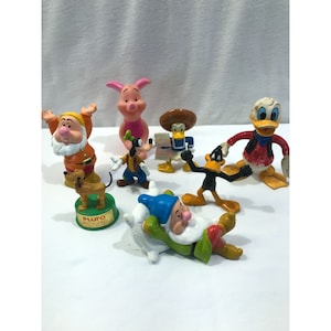 Könnte beinhalten: Eine Sammlung farbenfroher Zeichentrickfiguren, darunter Schneewittchens Zwerg, Pluto, Ferkel, Goofy, Donald Duck und Daffy Duck. Die Figuren sind aus Kunststoff und auf einer weißen Oberfläche angeordnet.