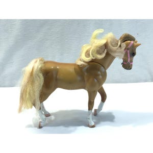 Pode incluir: Um cavalo de brinquedo de plástico castanho com crina e cauda loiras. O cavalo tem uma rédea rosa e marcas brancas nas pernas. O cavalo de brinquedo está sobre uma superfície branca.