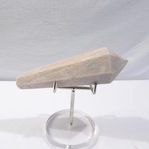 May include: A light beige crystal point, displayed on a silver metal stand with a clear acrylic base. The crystal has a faceted, pointed end and a smooth, textured surface. The stand elevates the crystal, showcasing its shape and color.