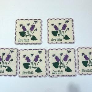 May include: Seven square coasters with scalloped edges. Each coaster features a purple violet floral design with green leaves and stems. The text "Devon Violets MADE IN ENGLAND" is printed on each coaster.