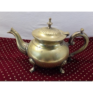 May include: A vintage brass teapot with a rounded body, a curved spout, and a decorative handle. The lid is adorned with a finial. The teapot rests on three small feet. The overall design suggests a classic, antique aesthetic.