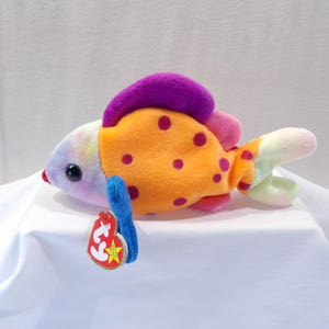 Puede incluir: Un juguete de peluche de pescado colorido con piel naranja, morada, rosa y blanca. El pez tiene un cuerpo grande y redondo con una cabeza pequeña y puntiaguda y una cola larga y delgada. El pez tiene una etiqueta azul unida a su lado con la palabra "Ty" en ella.