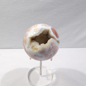 May include: A spherical geode with a polished exterior, showcasing a mix of white, peach, and reddish-brown hues. The geode is mounted on a clear, circular stand, revealing a hollow interior filled with crystalline formations. A decorative item.