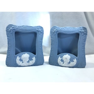 May include: A pair of light blue decorative frames with white cameo designs. The frames are rectangular with detailed edges. Each frame has a central opening and a white cameo depicting a woman's profile surrounded by roses.