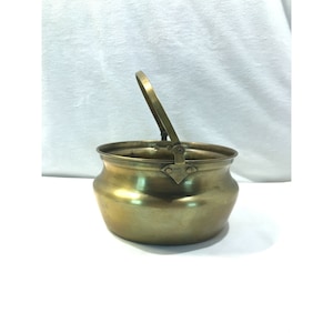 Puede incluir: Un pequeño cubo de latón antiguo con un asa curva. El cubo tiene un cuerpo redondeado y un borde acampanado. El asa está unida al cubo con bisagras. El latón tiene un color dorado cálido.